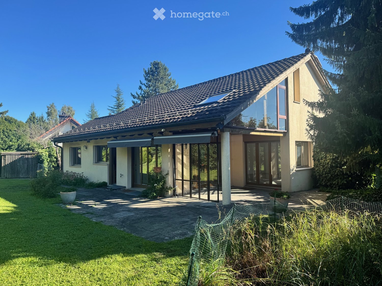 Two-story house with a modern design, featuring a tiled roof, large windows, and a covered entrance area. The surrounding yard includes a well-maintained lawn, a paved pathway, and some potted plants.