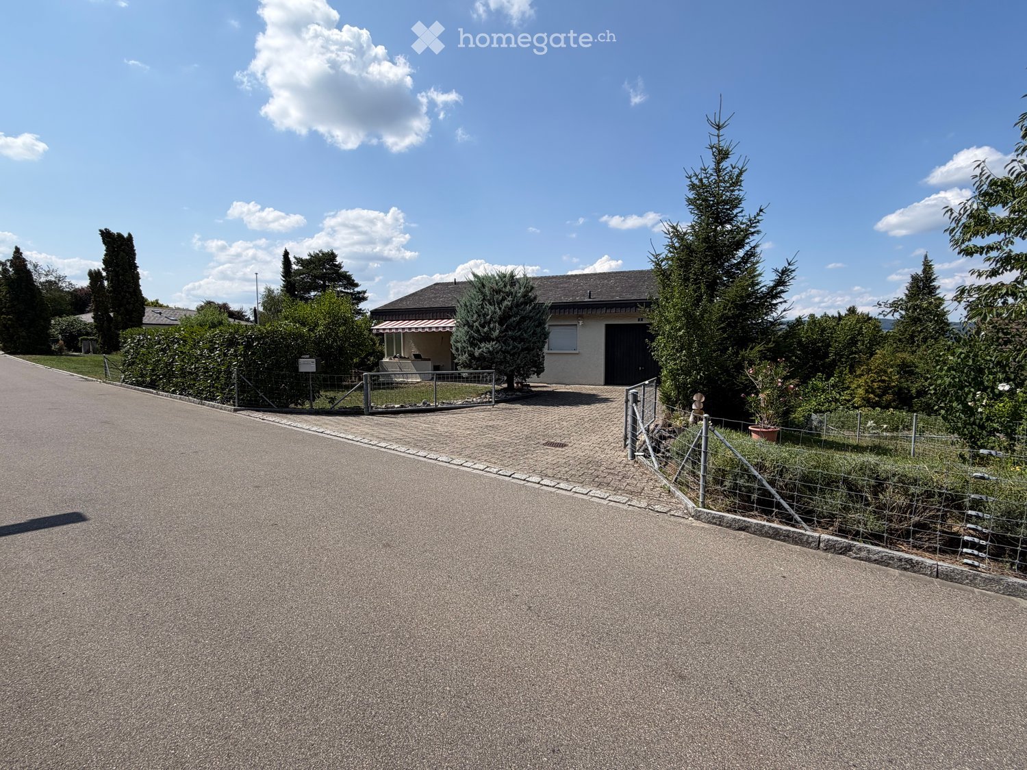 Single-story house with a dark garage door, surrounded by trees and shrubs, with a paved driveway and a metal fence.
