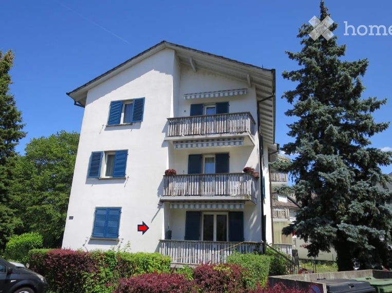 A three-story white building with blue shutters and balconies, surrounded by greenery and trees.