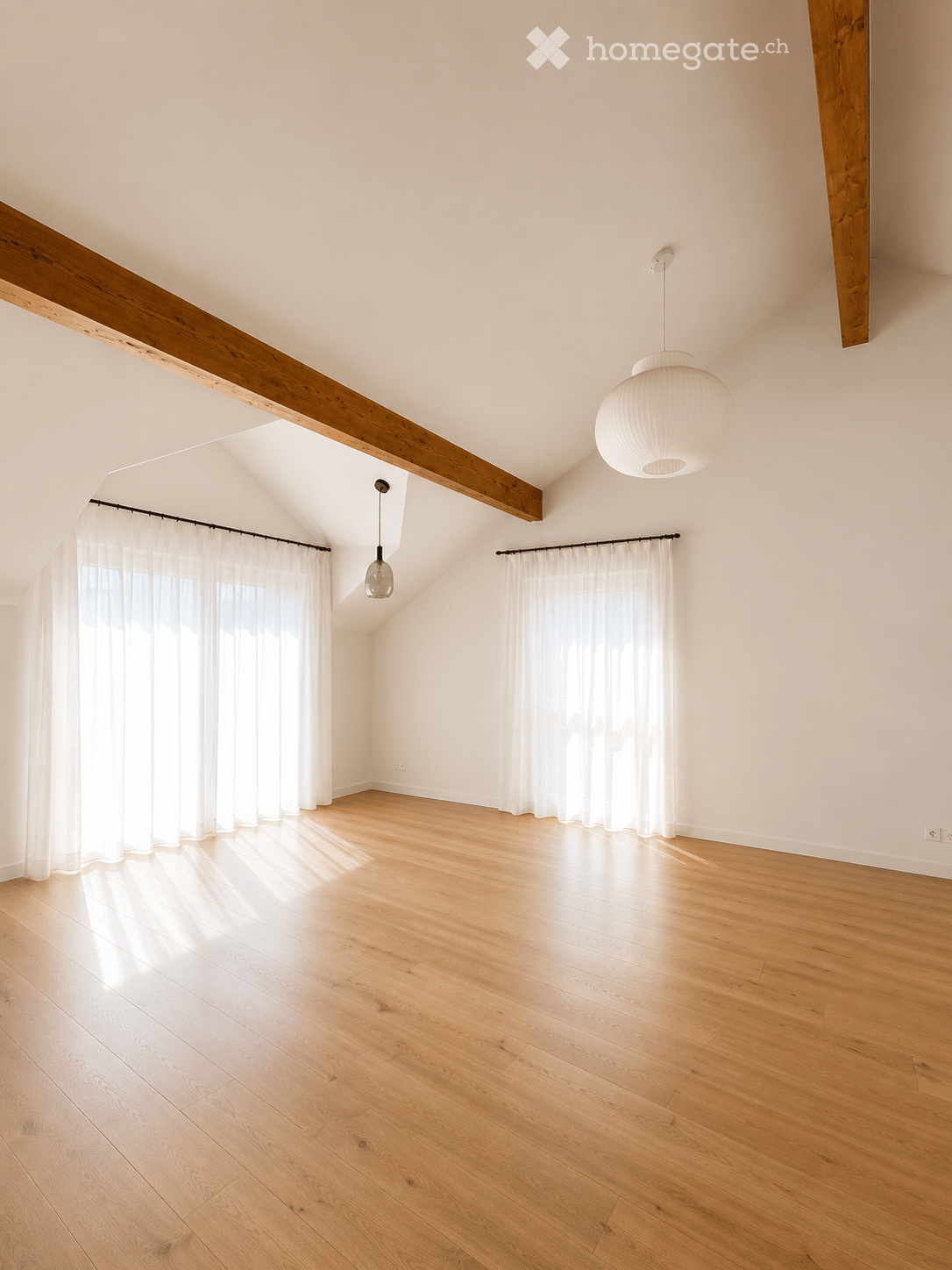 Bright, empty room with wooden flooring, white walls, large windows with white curtains, and exposed wooden beams on the ceiling.