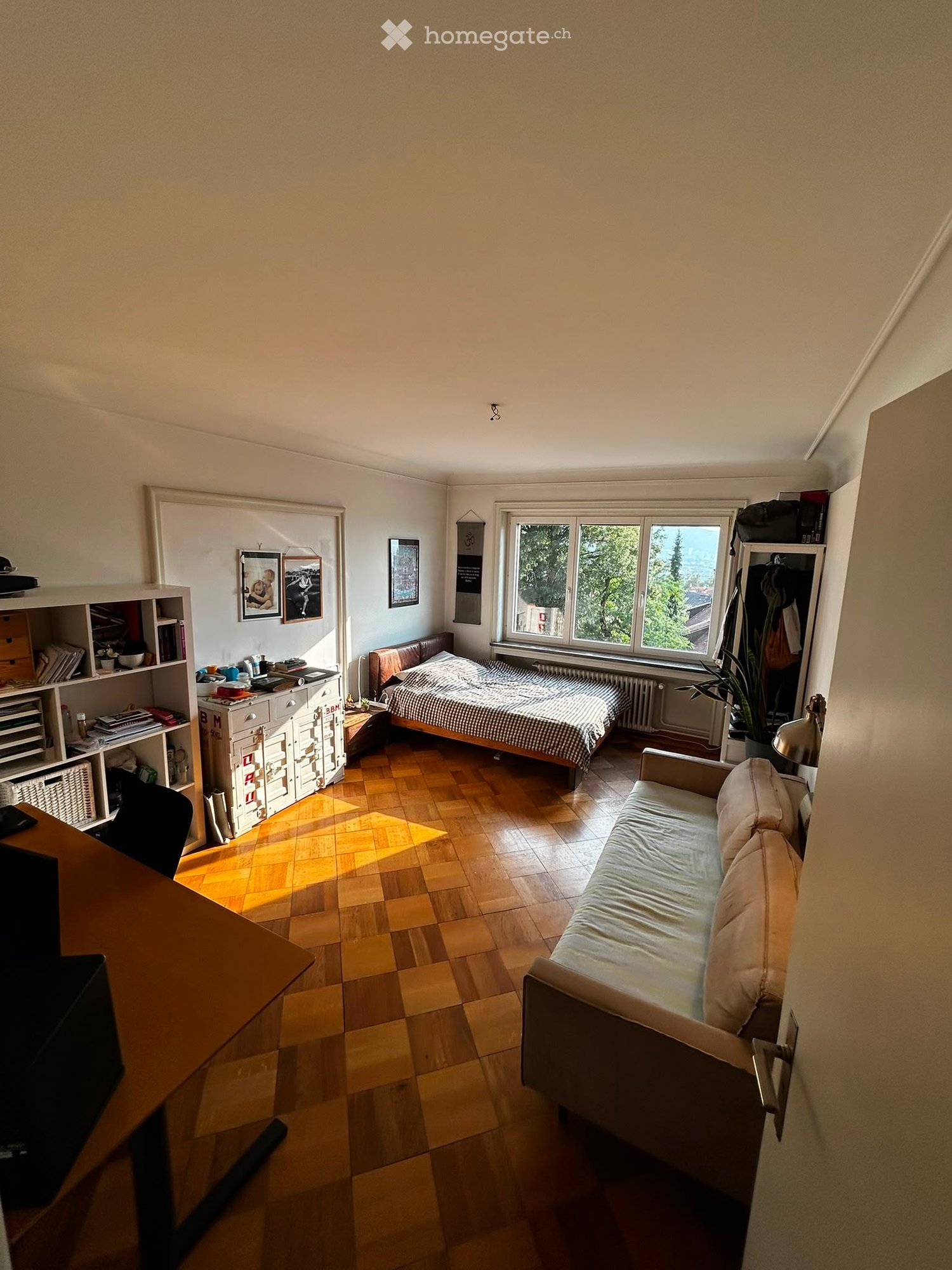 A well-lit bedroom with wooden flooring, a bed with a checkered duvet, a sofa, a desk with a chair, a cabinet, and a potted plant near the window.
