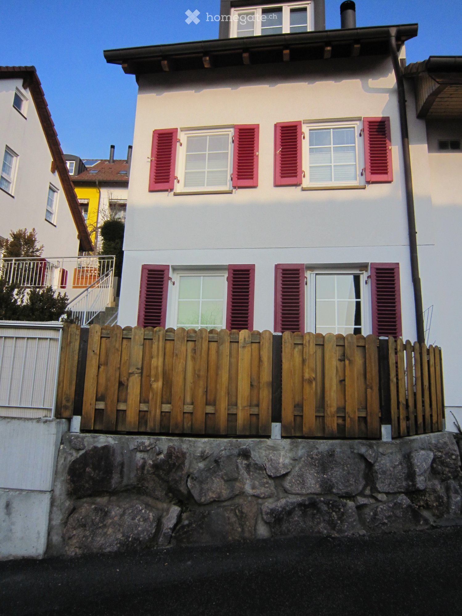 Two-story house with white walls, red shutters, and a wooden fence on a raised ground floor.