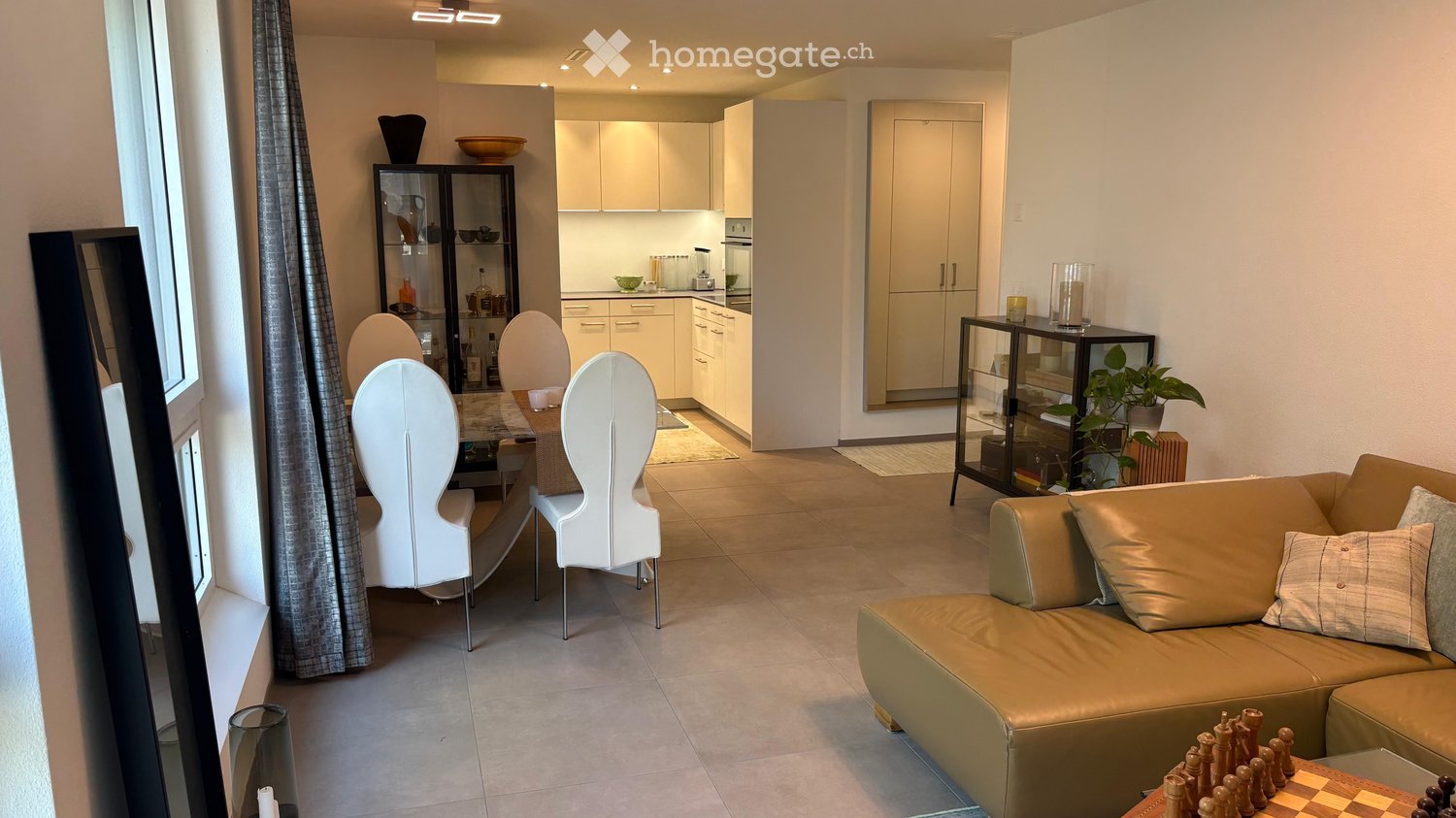 Modern living and dining area with a beige sofa, white chairs, a glass coffee table, and a dining table with four white chairs. The kitchen features white cabinets and a countertop with various appliances.