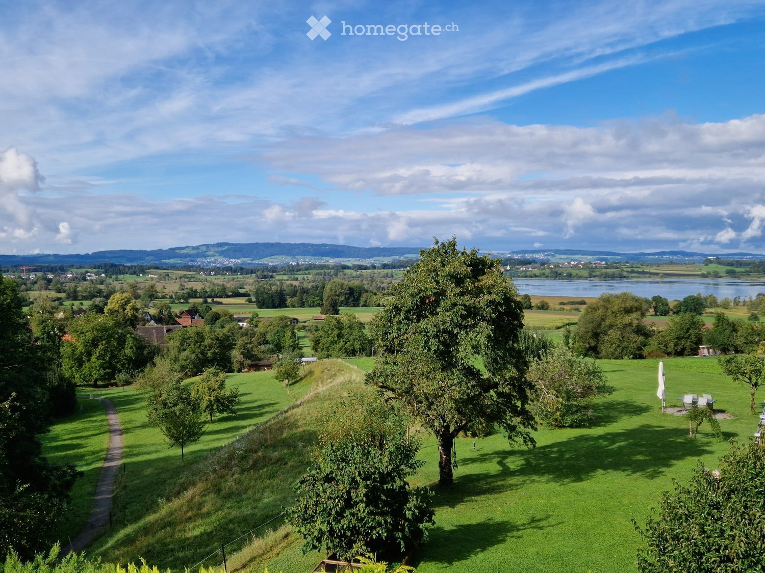 A panoramic view of a lush green garden with trees, a pathway, and a scenic landscape featuring fields, a lake, and distant hills under a partly cloudy sky.