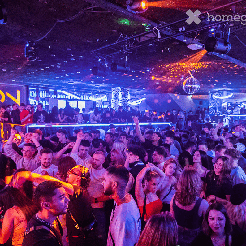 Large crowd inside a club with purple and red lights, disco ball, elevated bar area with bottles, dance floor.