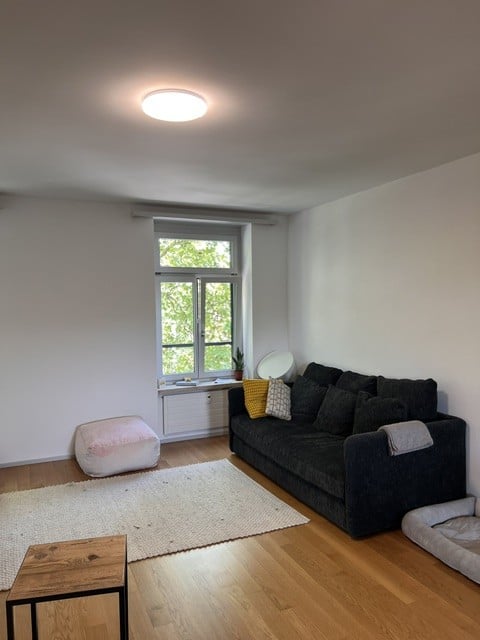 Modern living room with wooden flooring, white walls, large window, dark grey sofa, ottoman, coffee table, and floor lamp.
