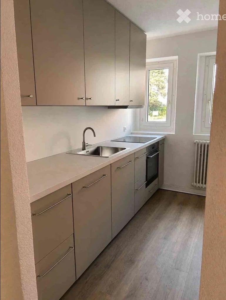 Modern kitchen with light grey cabinets, a stainless steel sink, and a built-in oven. The space features light wooden flooring and large windows allowing natural light.
