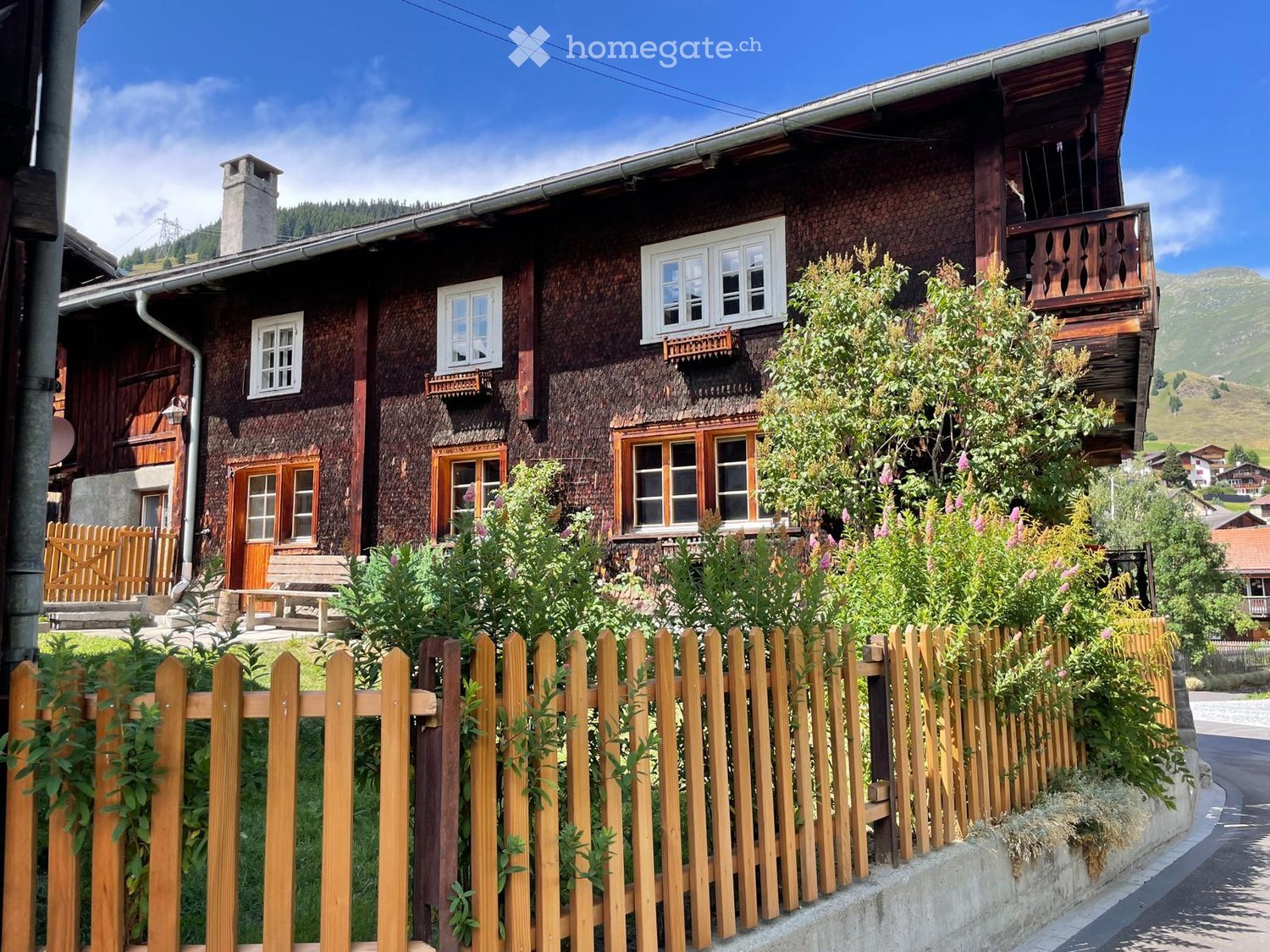 Traditional wooden house with multiple windows, a balcony, and a wooden fence in front, surrounded by greenery and a scenic mountain backdrop.