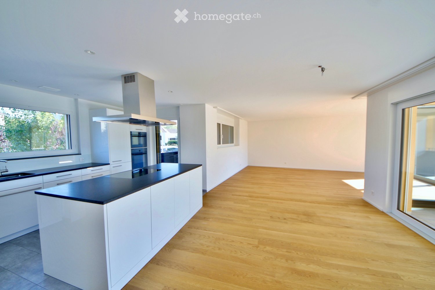 Modern kitchen with white cabinets, black countertops, a central island, and built-in appliances including a sink, oven, and range hood. The space features light wooden flooring and large windows allowing natural light.