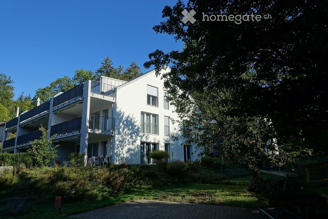 Modern white multi-story building with balconies, surrounded by greenery and trees.