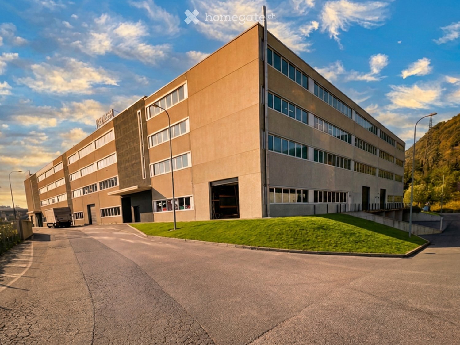 Multi-story industrial building with large windows, beige facade, and a visible garage entrance. The building is surrounded by a paved area and a small patch of grass.