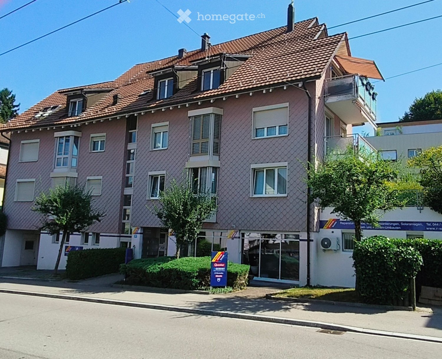 Two-story residential building with pink facade, multiple windows, and a tiled roof. Features a balcony on the upper floor and a small garden area in front.