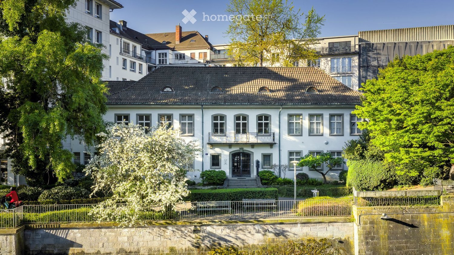 A large, white, multi-story house with a pitched roof, multiple windows, and a small balcony. The house is surrounded by greenery and trees, with a well-maintained garden in front.