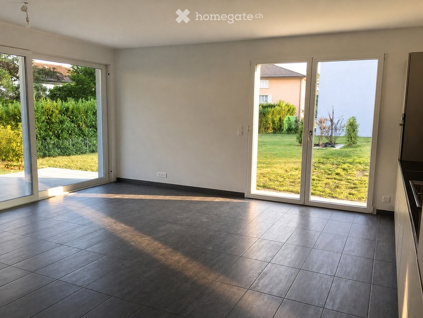 Empty room with large sliding glass doors leading to a garden, tiled floor, and white walls.