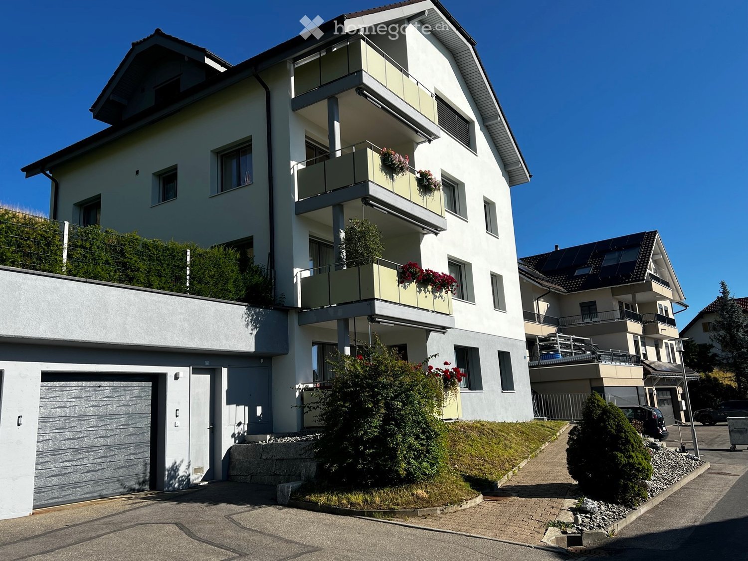 Modern two-story house with white exterior, multiple balconies, garage, and surrounding greenery.