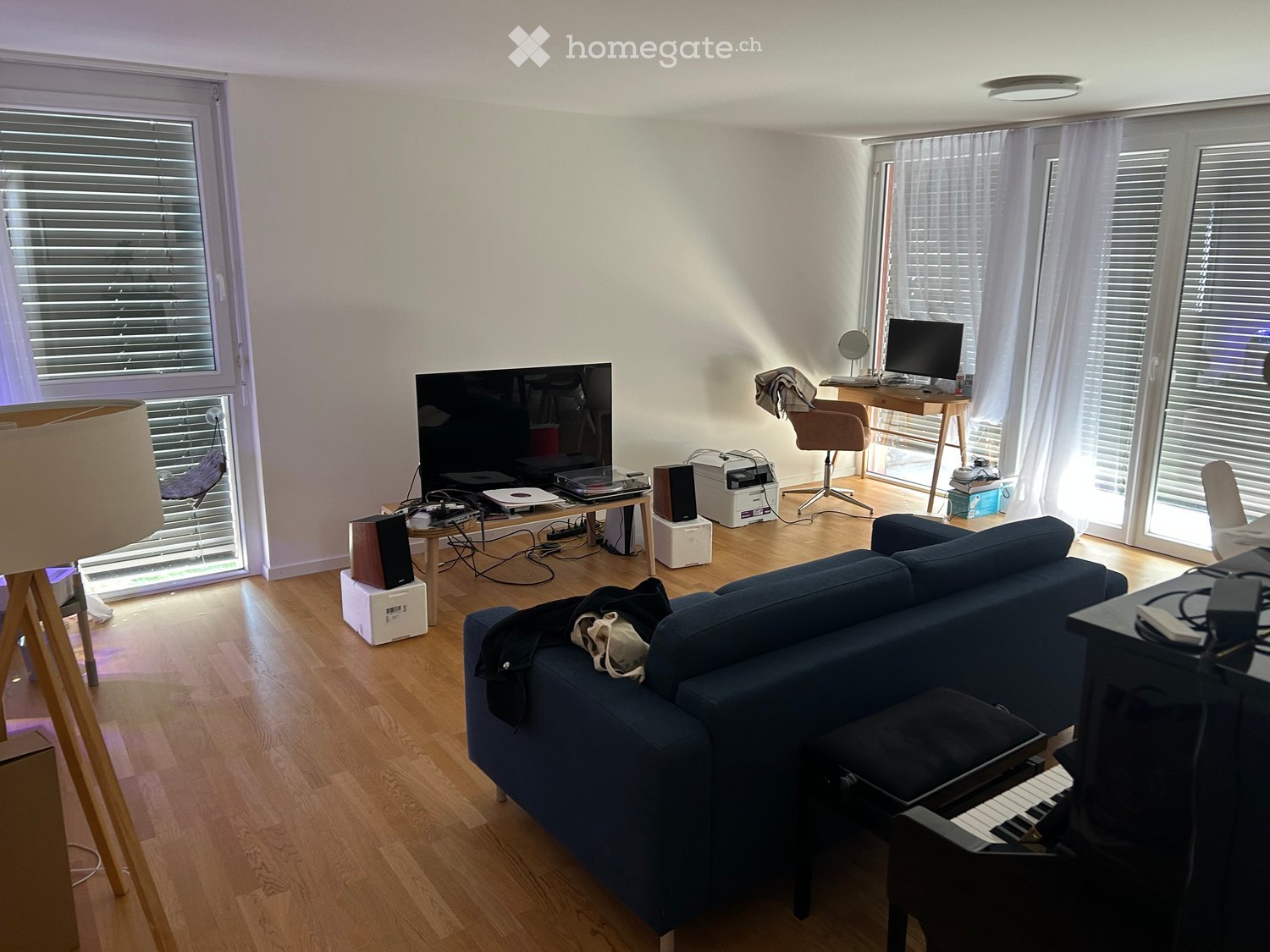 Living room with wooden flooring, large windows, a blue sofa, a piano, a table with a screen, and a chair.