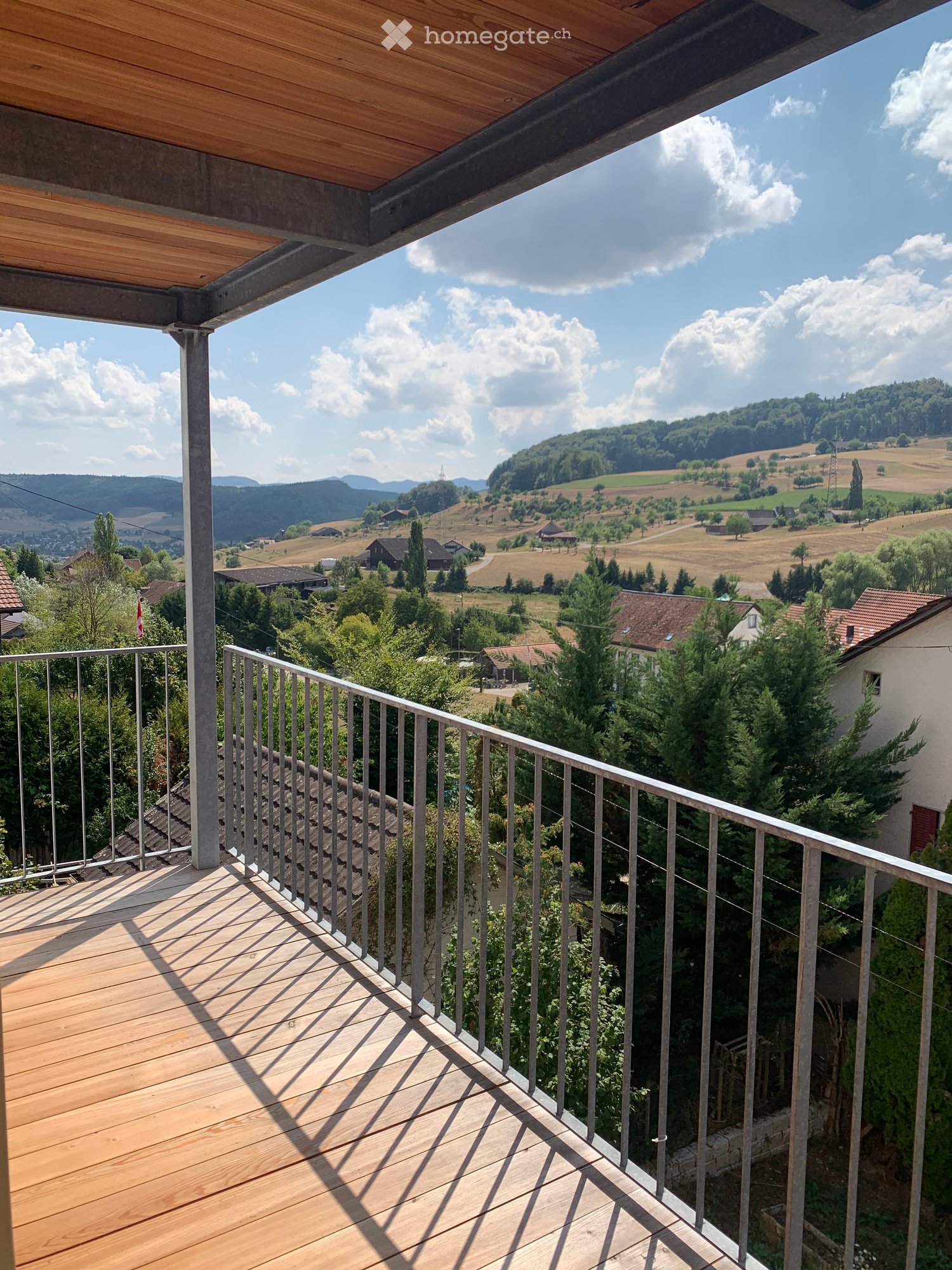 Wooden balcony with metal railing, overlooking a scenic countryside with fields, trees, and distant hills.