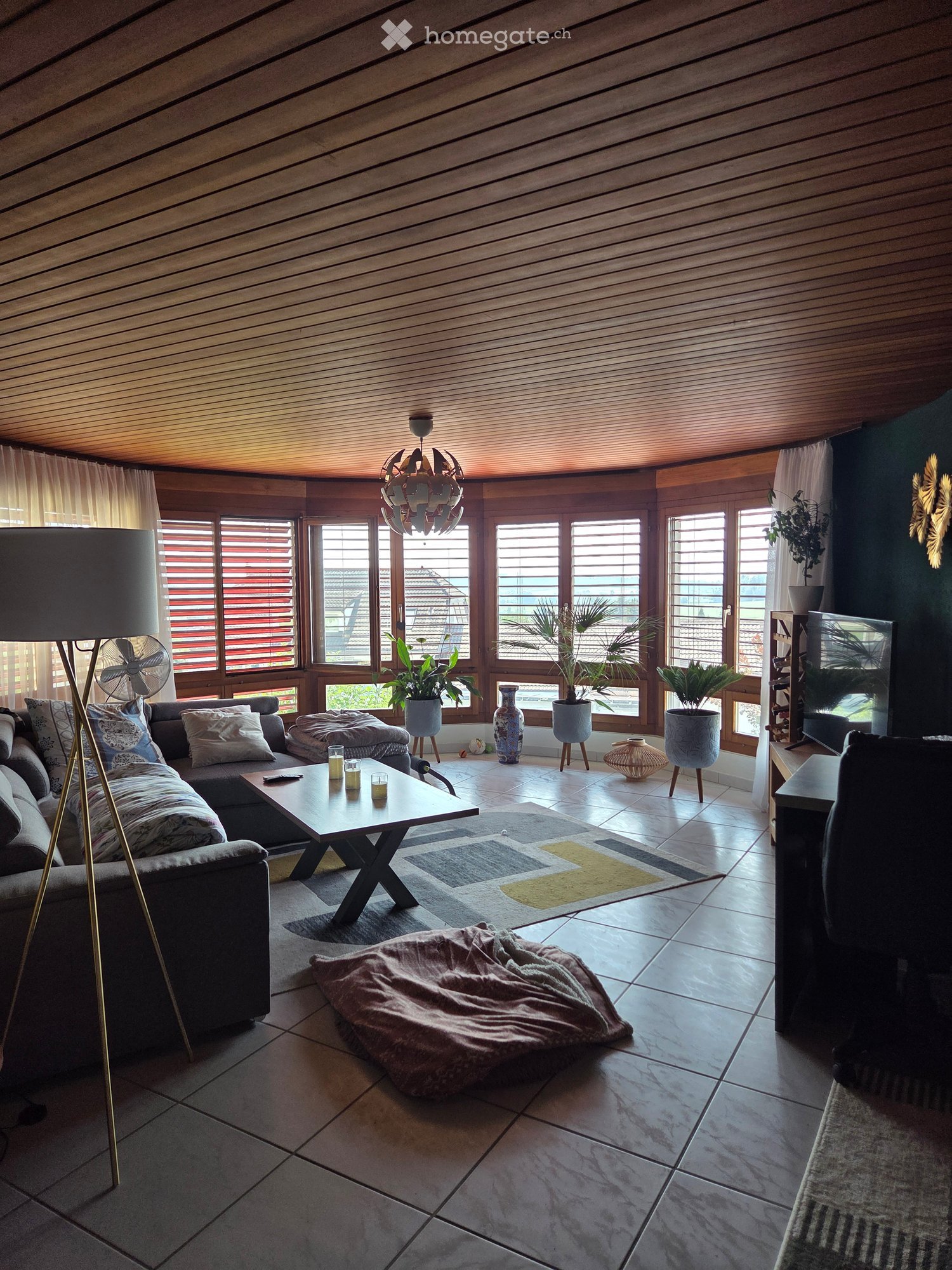 Modern living room with large windows, wooden ceiling, grey sofa, coffee table, floor lamp, fan, and decorative vases.