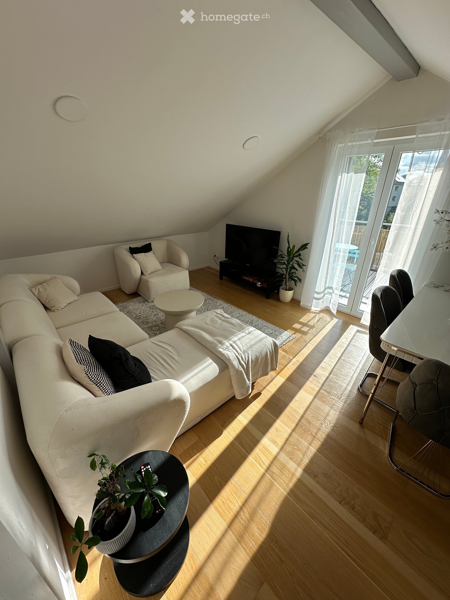 Bright living room with wooden flooring, large windows, and a balcony door. Features a white sectional sofa, armchair, coffee table, TV, and dining area with chairs and table.