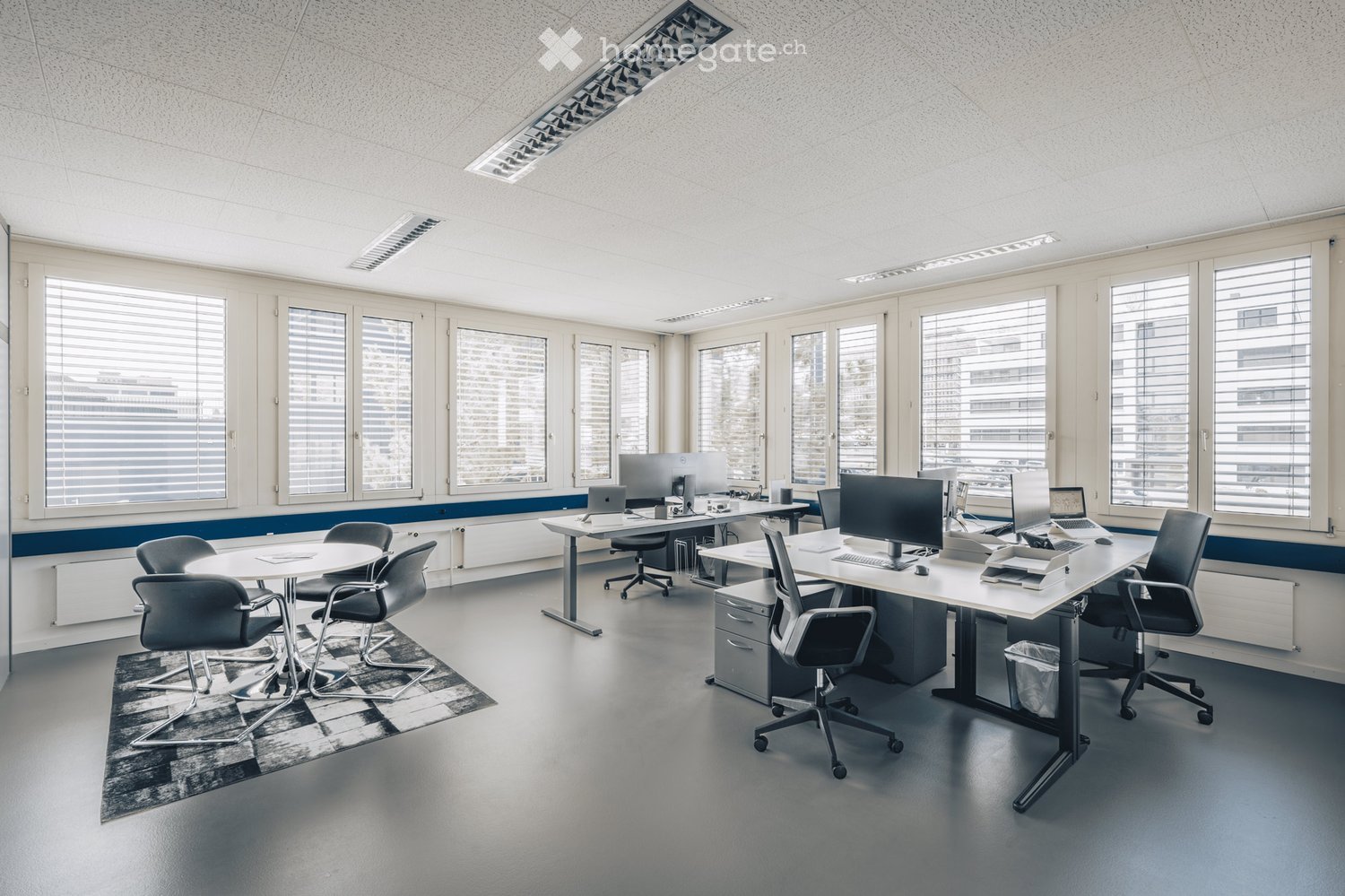 Modern office space with multiple desks, chairs, laptops, computers, and a printer. Large windows with blinds provide natural light.