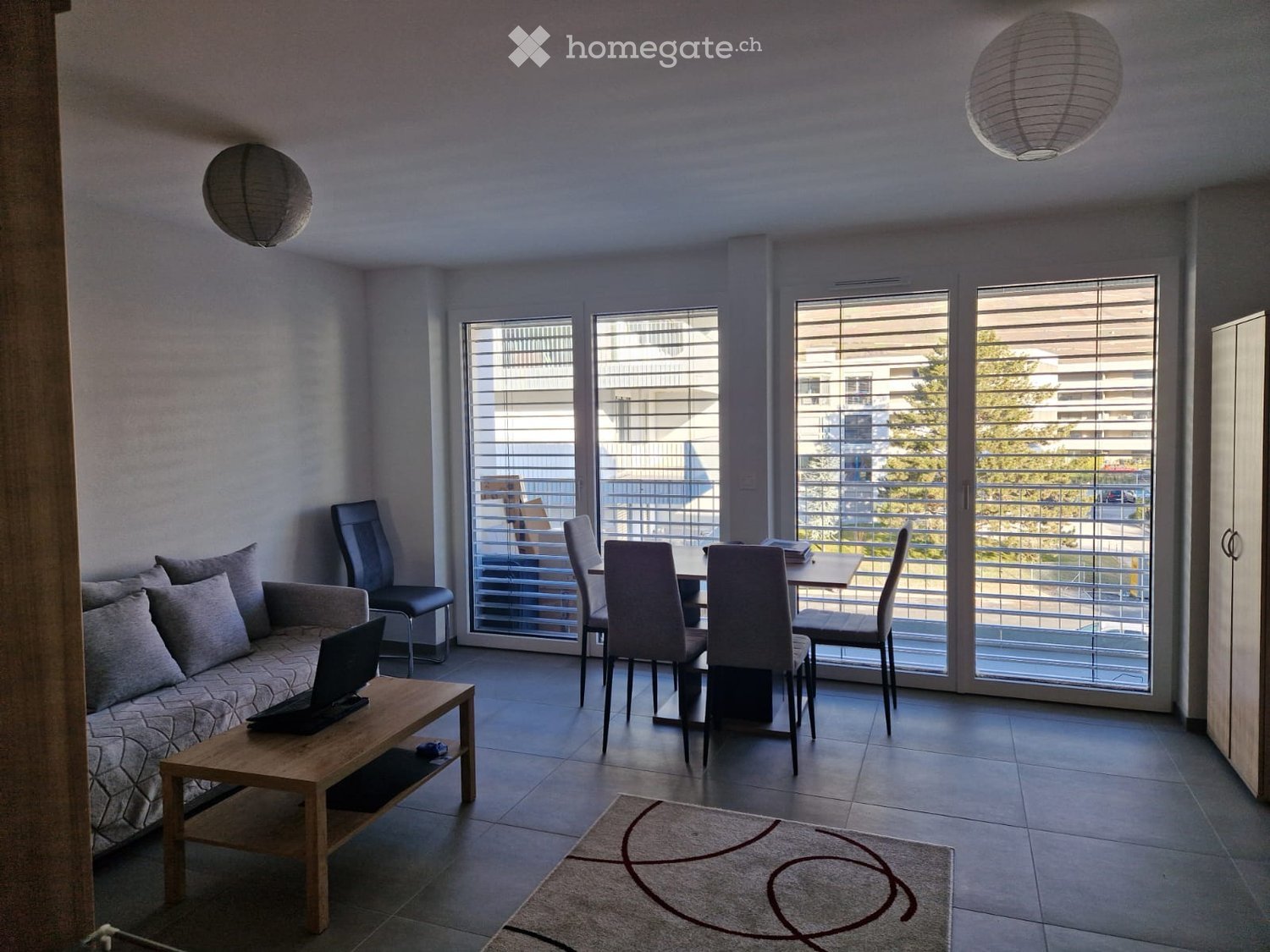 Modern living and dining area with a sofa, coffee table, laptop, dining table, chairs, and pendant lamps. Large windows with blinds lead to a balcony.