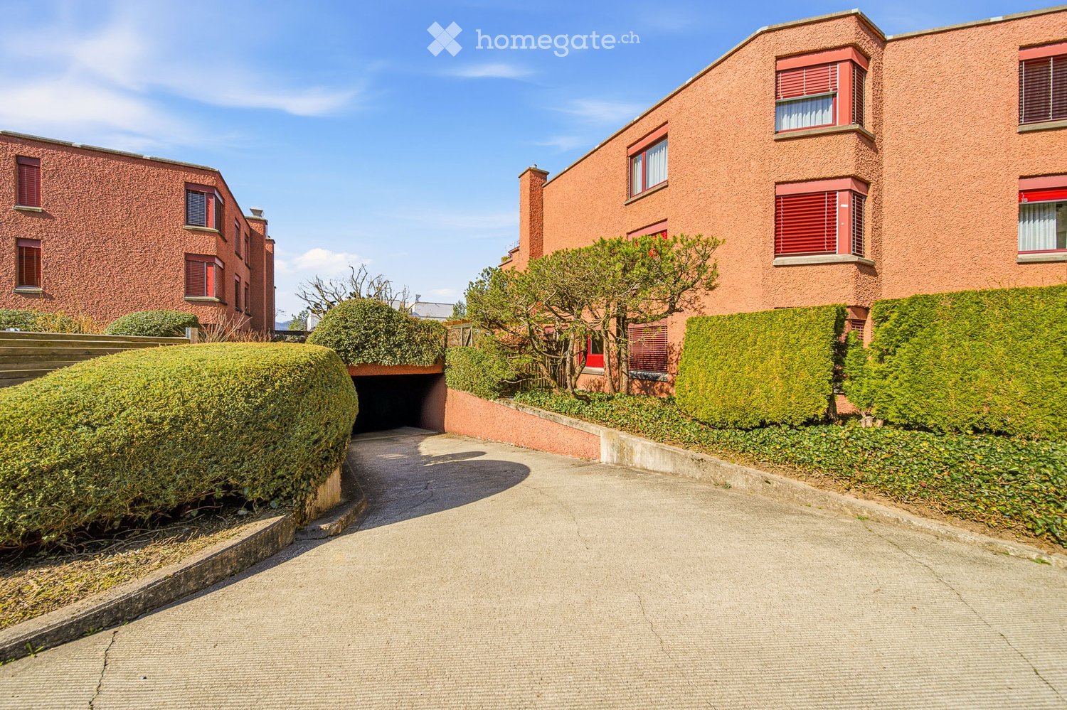 Two-story red brick building with multiple windows and a raised ground floor entrance leading to a garage. The building is surrounded by greenery and hedges.
