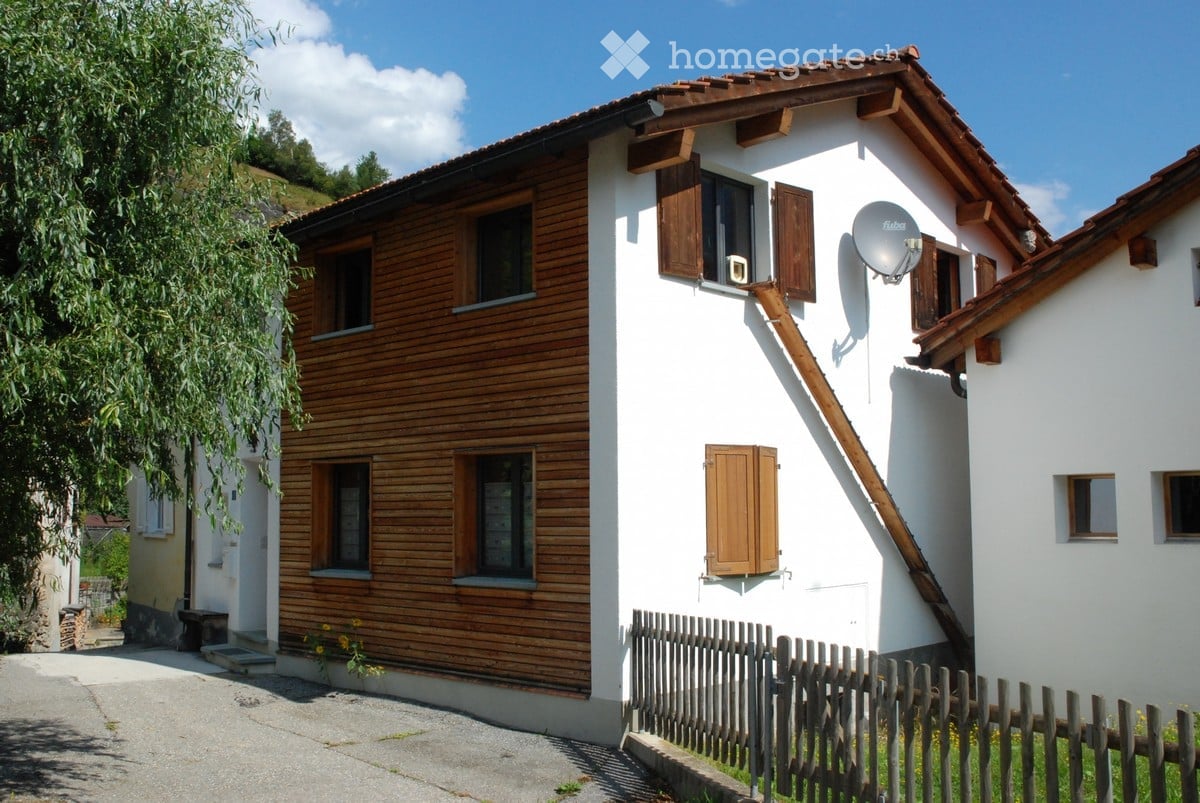 Two-story house with white walls, wooden shutters, and a satellite dish on the roof. The house features a wooden fence and is surrounded by greenery.