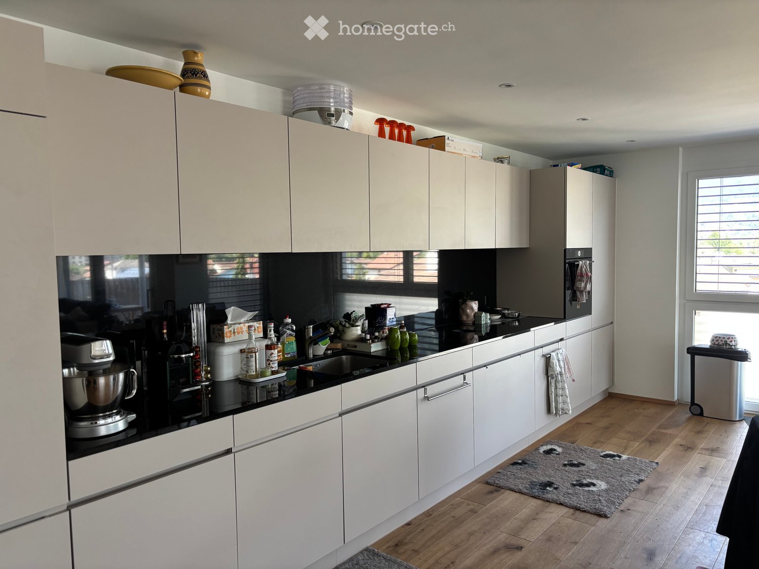 Modern kitchen with white cabinets, black countertops, and wooden flooring. Visible appliances include a mixer, coffee maker, and oven. Bottles and cups are on the counter, and a rug is on the floor.