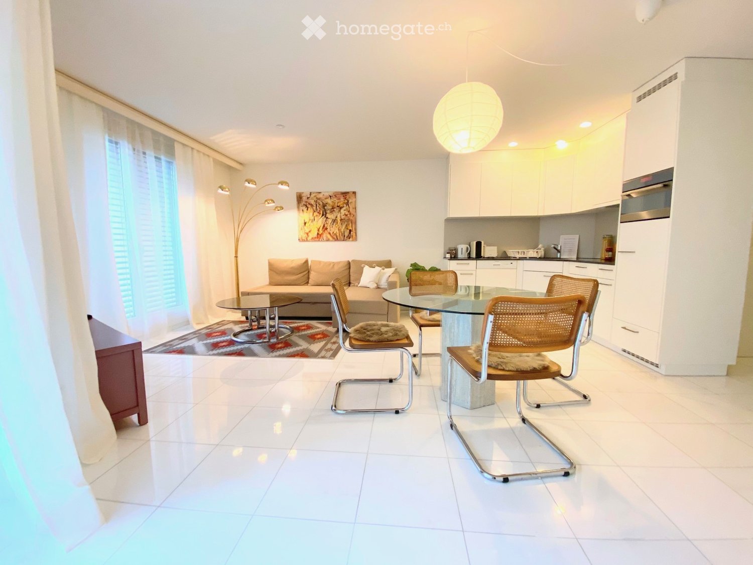 Modern living and dining area with white tiled floors, a sofa, coffee table, dining table with chairs, kitchen area with cabinets and appliances, and a floor lamp.