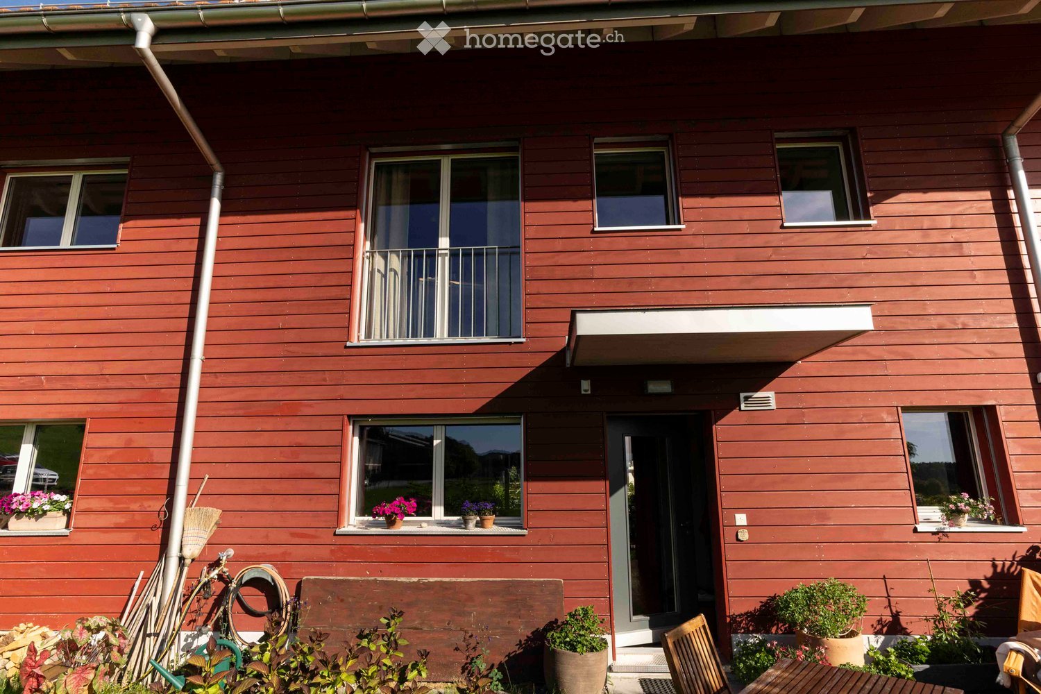 Two-story wooden house with red siding, multiple windows, and a balcony on the upper floor. The front yard features potted plants, a wooden bench, and a small table with chairs.