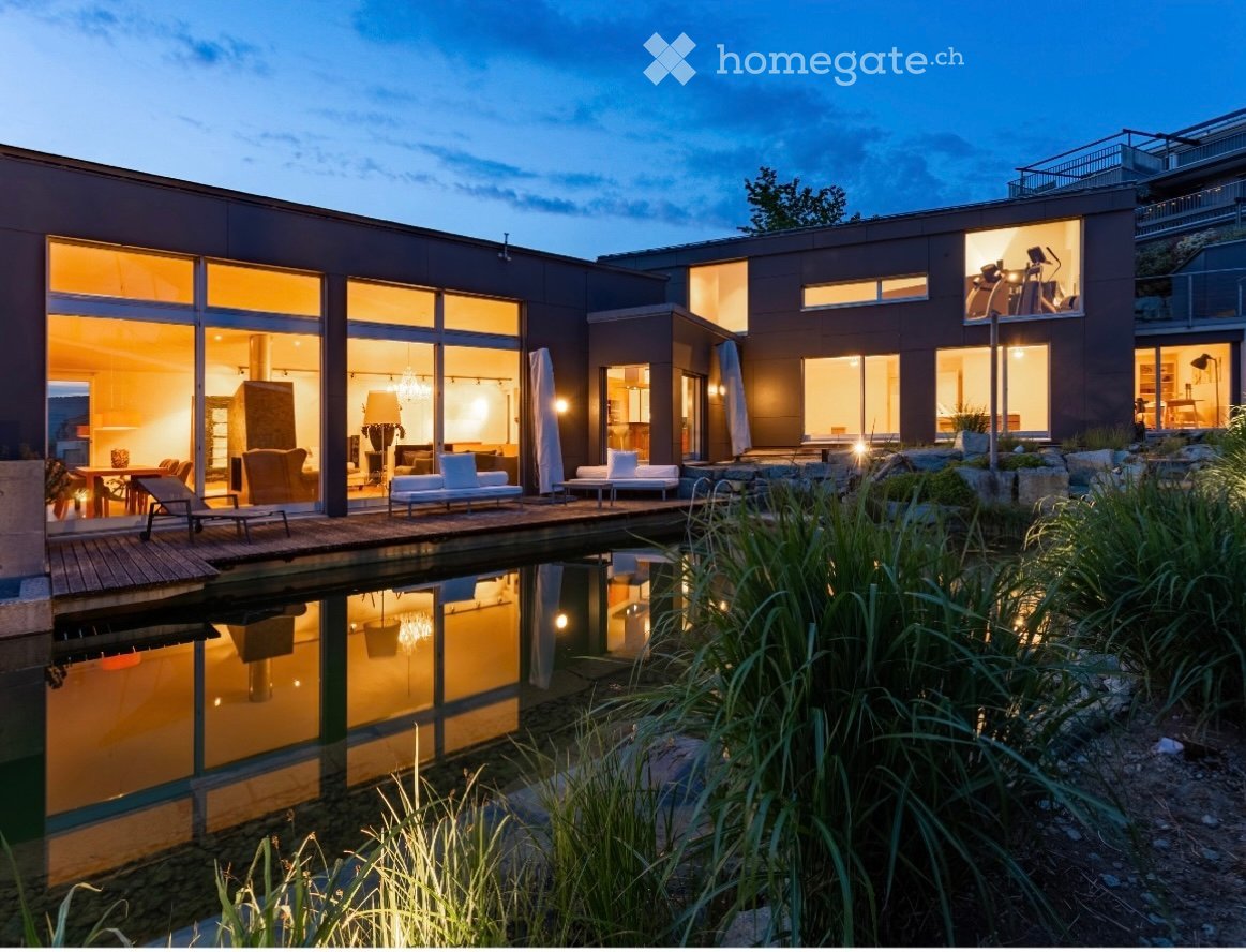 Modern two-story house with large glass windows, a reflective water feature, and outdoor seating area.