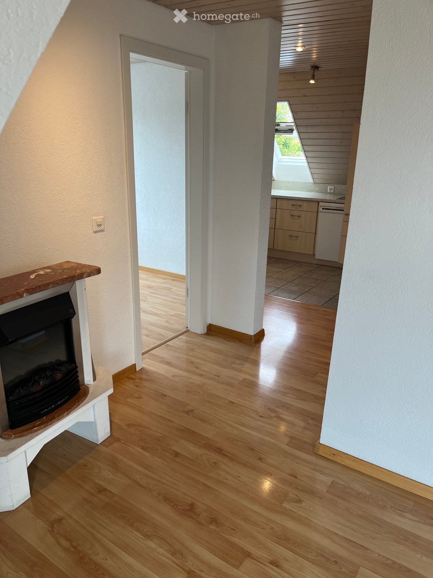 Spacious hallway with wooden flooring, white walls, and a fireplace on the left side. An open doorway leads to another room with light-colored cabinets and a tiled floor.