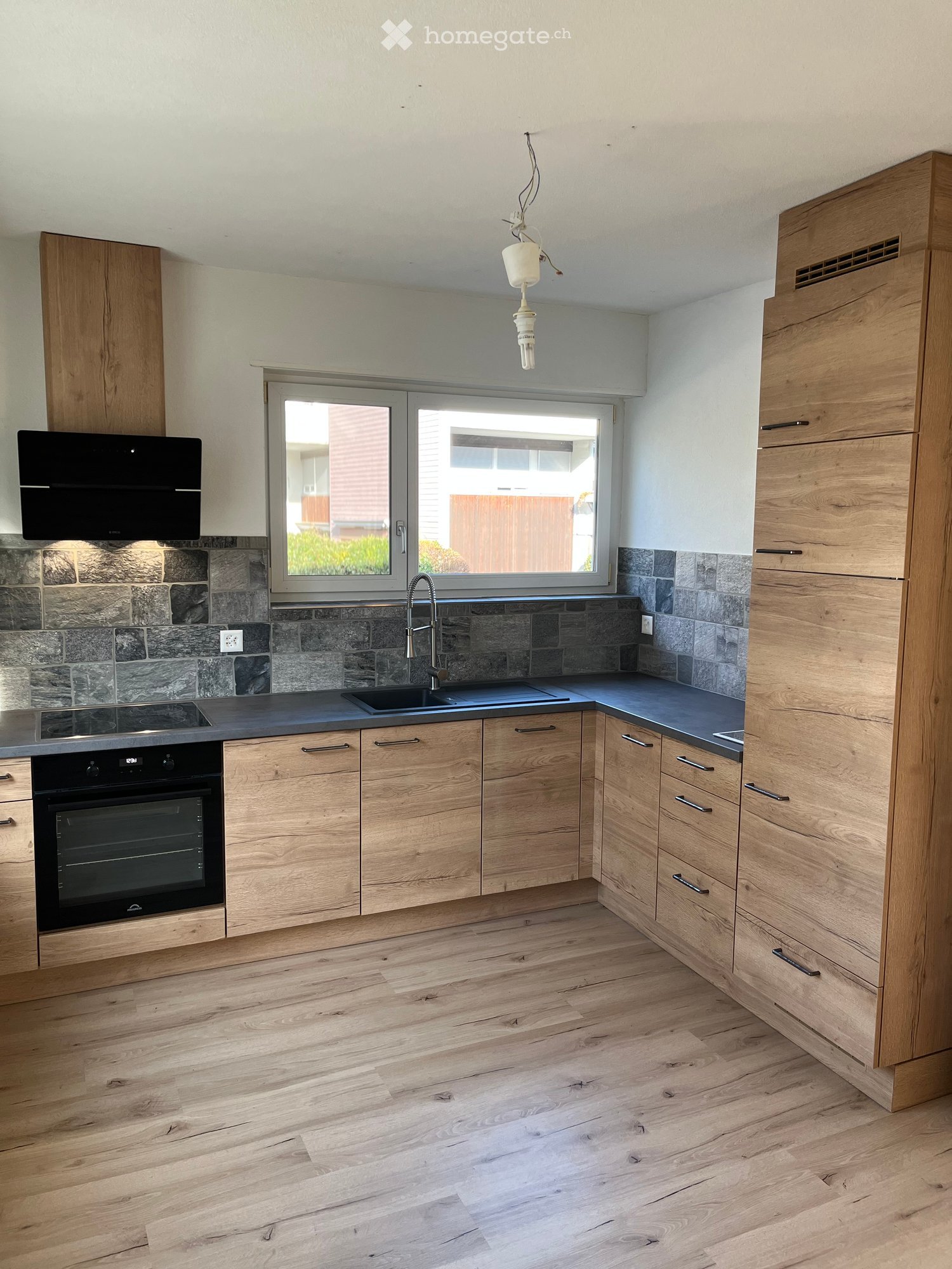 Modern kitchen with light wooden cabinets, dark countertops, a sink, and an oven. The backsplash is made of stone tiles, and there is a window above the sink.