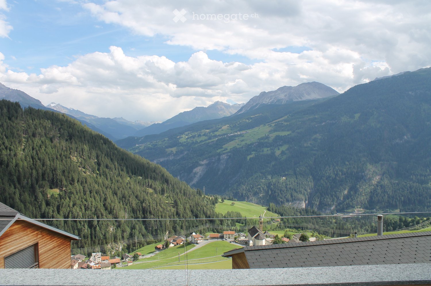 A panoramic view from a balcony or terrace overlooking a lush green valley with scattered houses, surrounded by dense forests and towering mountains under a partly cloudy sky.