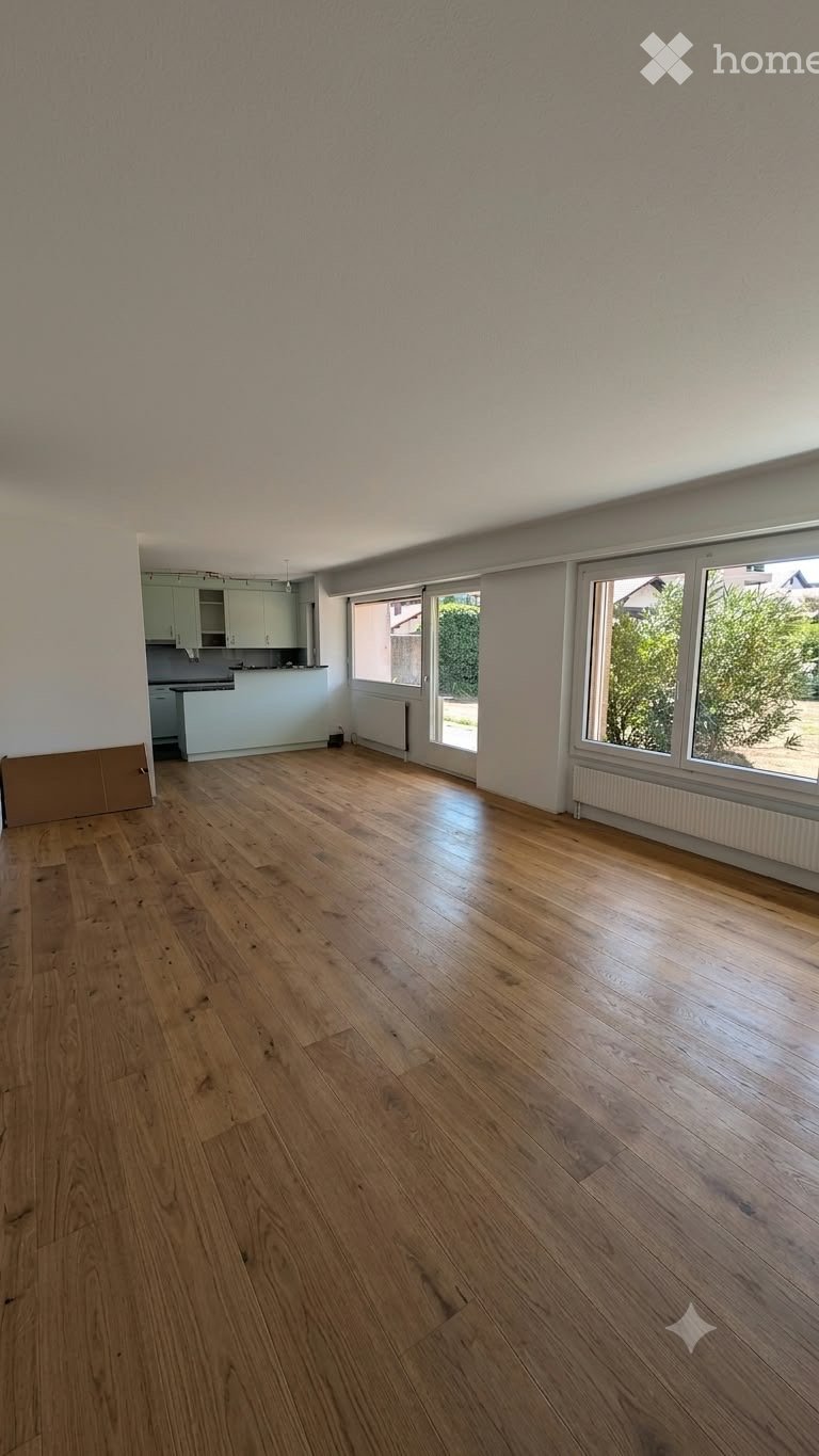 Empty room with light wooden flooring, white walls, and large windows. The adjacent kitchen area is visible with white cabinets and a countertop.