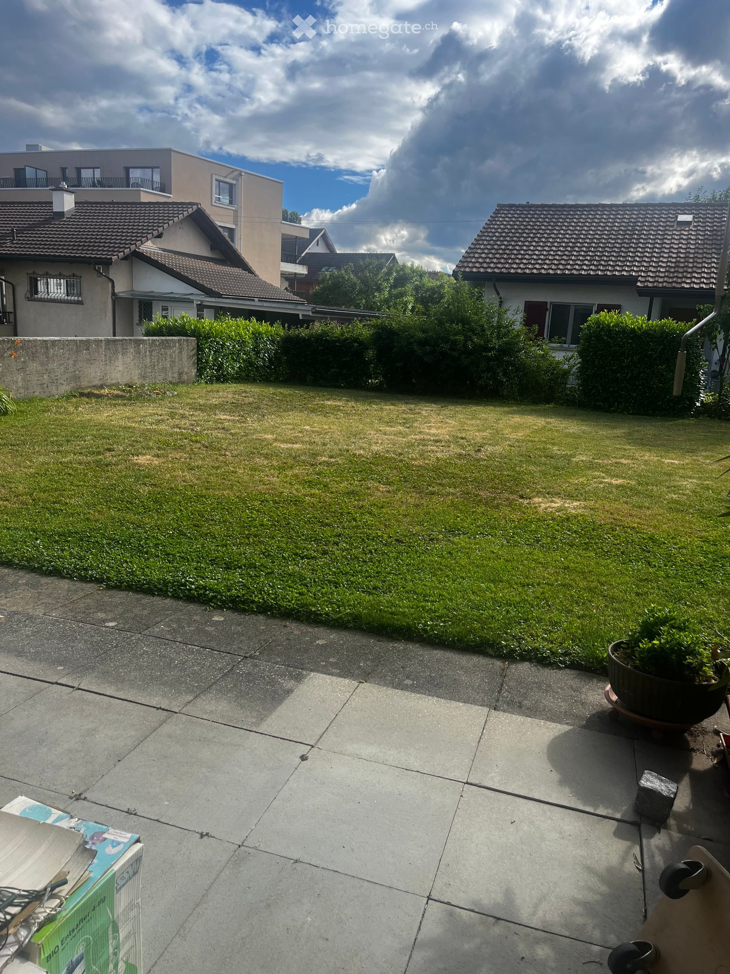 A well-maintained garden with a lawn, trimmed hedges, and a concrete patio area. There are several houses visible in the background, and the sky is partly cloudy.