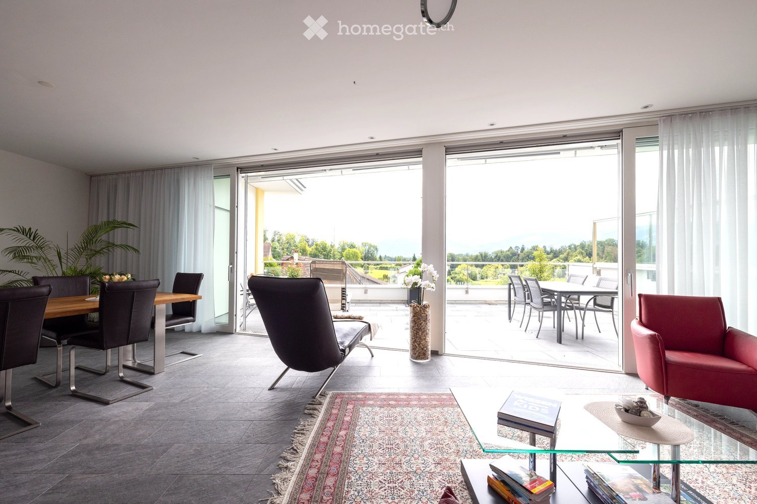 Modern living and dining area with large sliding glass doors opening to a balcony, featuring a red leather armchair, glass coffee table, dining table with chairs, and decorative vases.