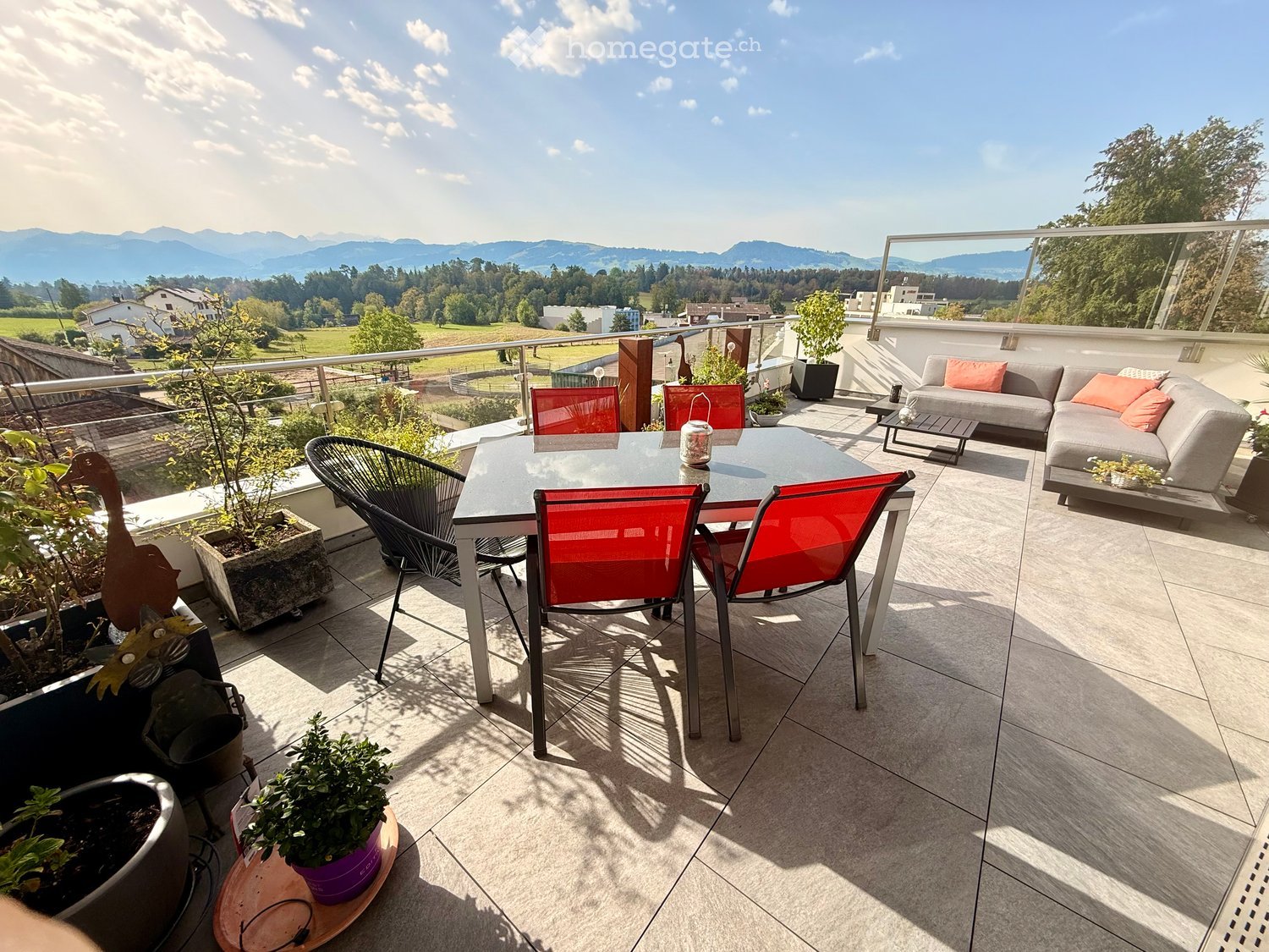 Balcony with a dining table, red chairs, sofa, and potted plants, overlooking a scenic landscape.
