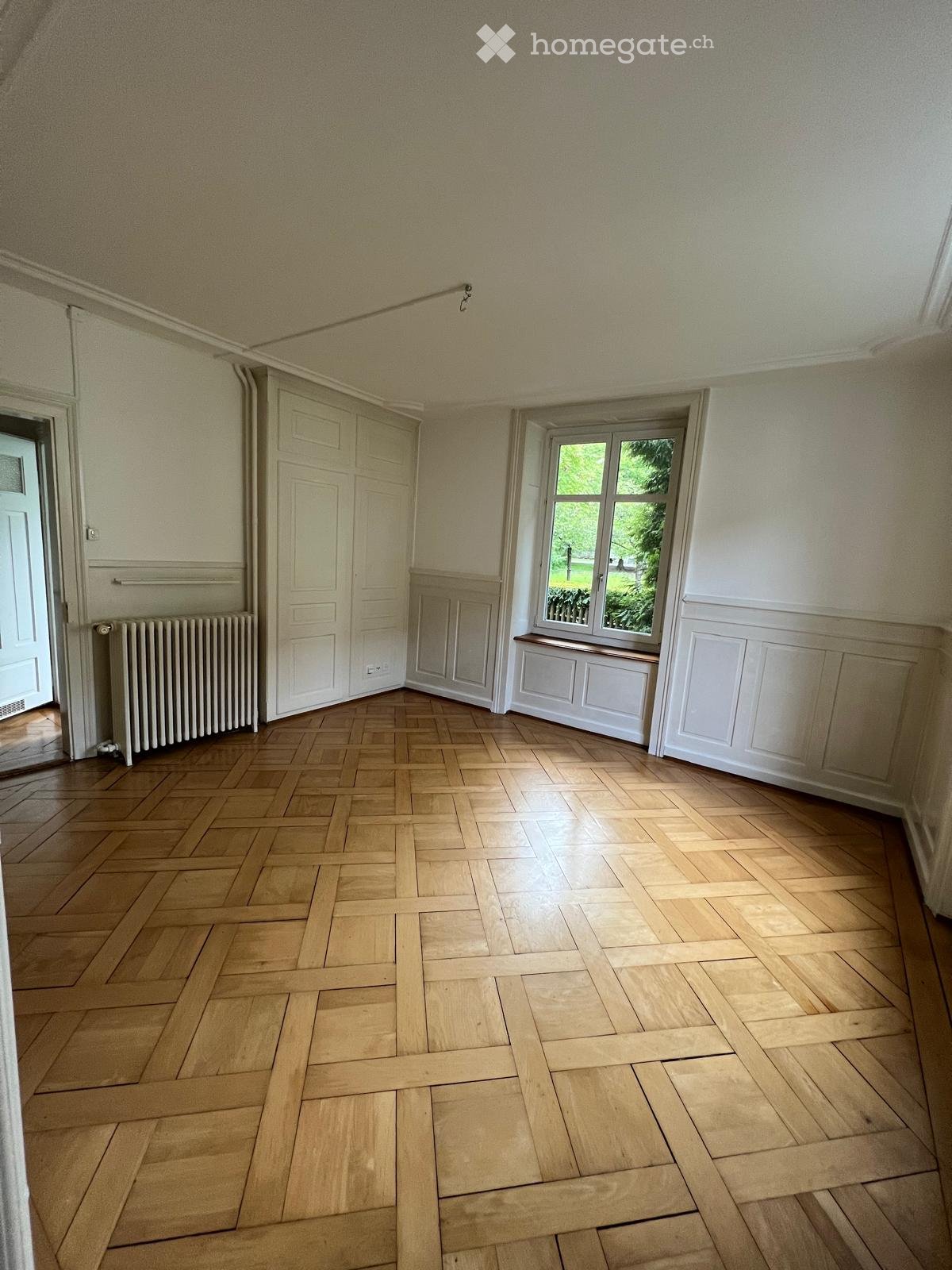 Empty room with wooden herringbone flooring, white walls, a window with a view of greenery, a radiator, and a closed door.