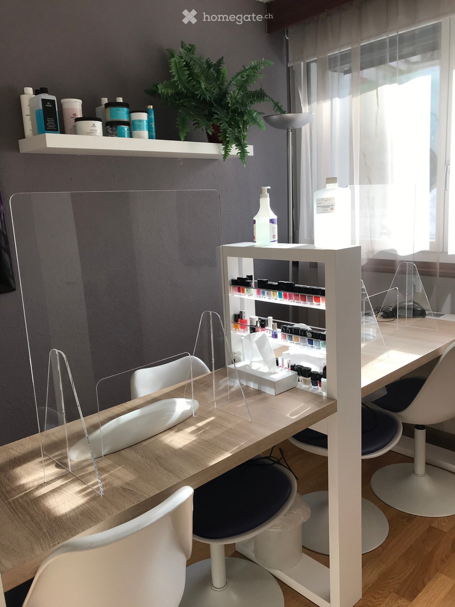 A modern, well-lit room with a wooden table, white chairs, shelves holding bottles and a plant, and a mirror on a stand.