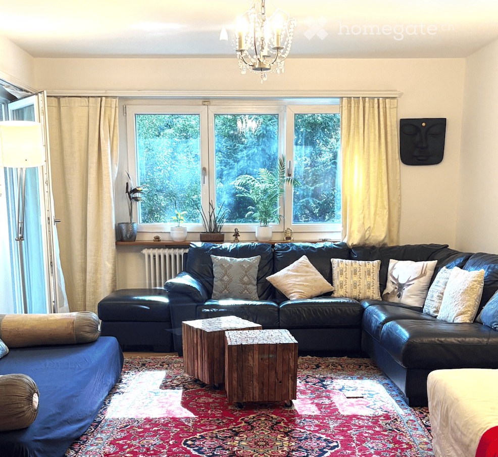 Modern living room with large windows offering a view, a black leather sectional sofa with multiple throw pillows, a wooden coffee table, a chandelier, and a patterned rug.