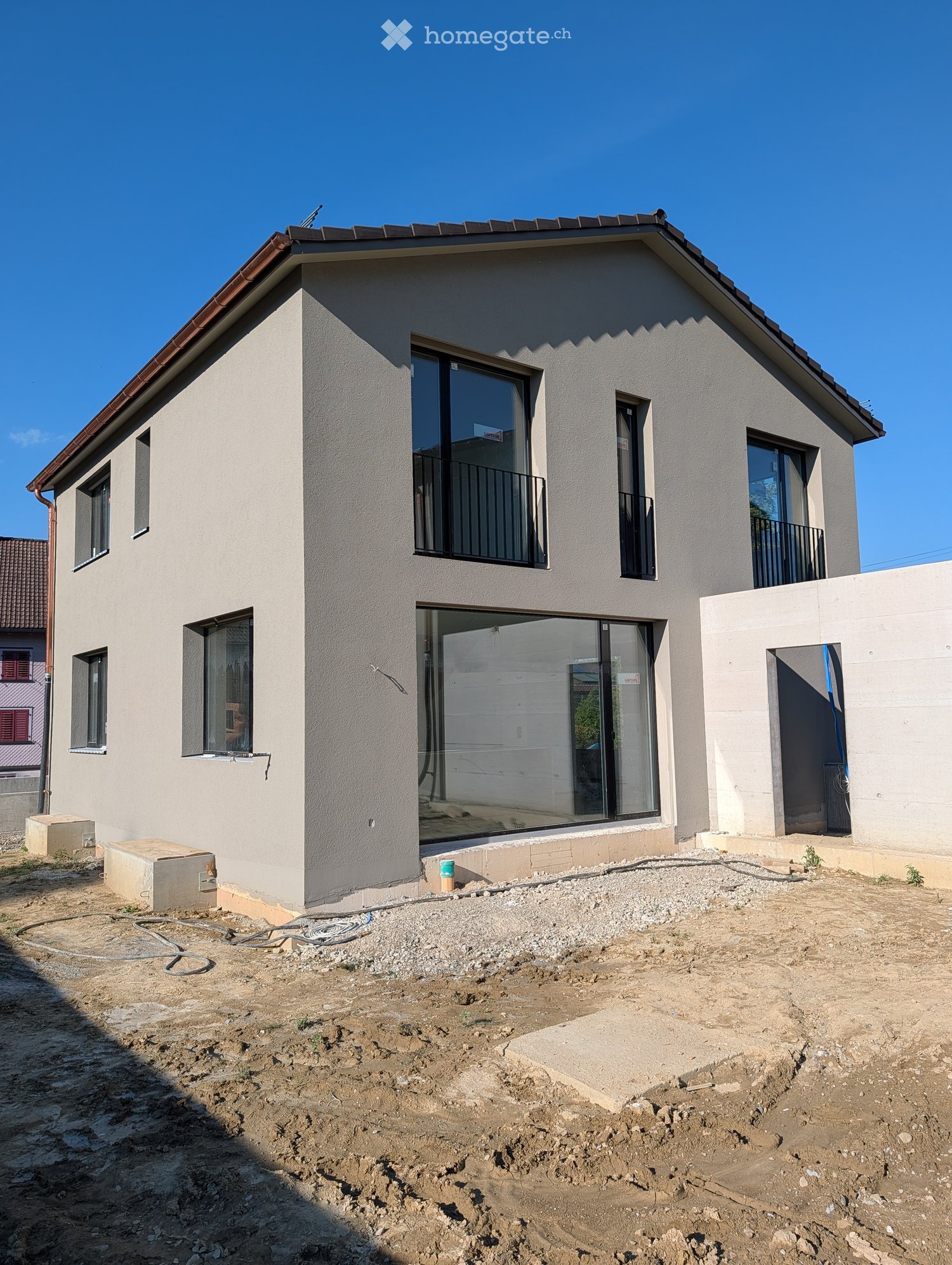 Two-story house with light gray exterior, large windows, and balconies. The ground floor has a raised entrance and unfinished yard with gravel and construction materials.