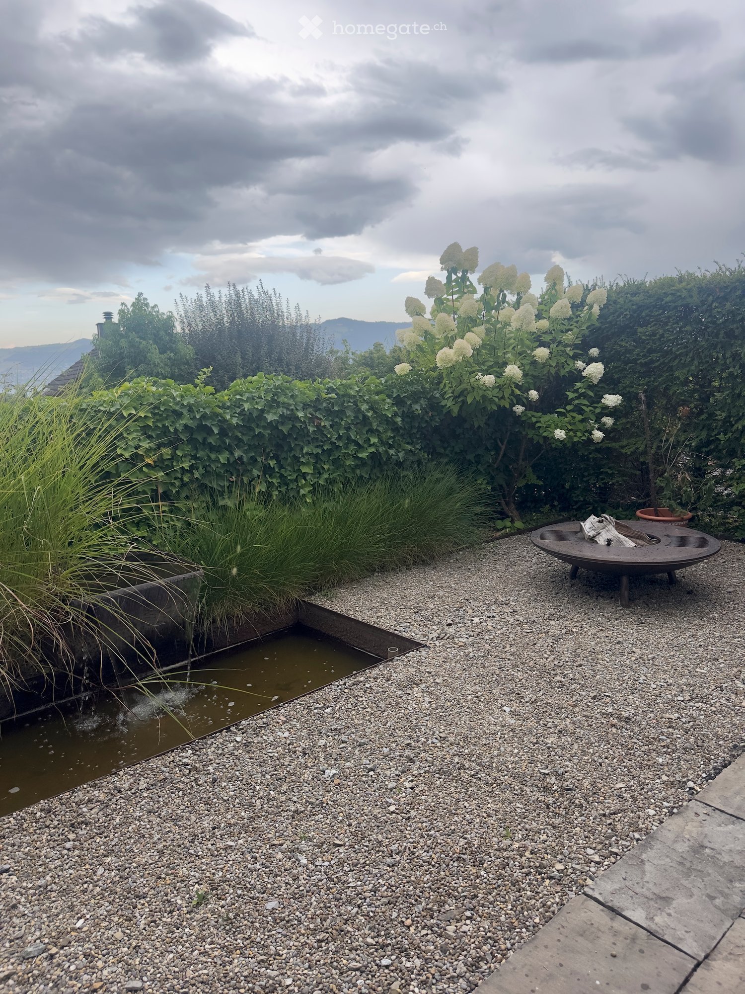 Garden area with gravel ground, small water feature, greenery, and a round metal table.