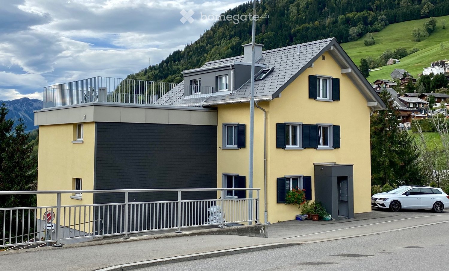 Two-story yellow house with grey roof and garage, surrounded by greenery and mountains.