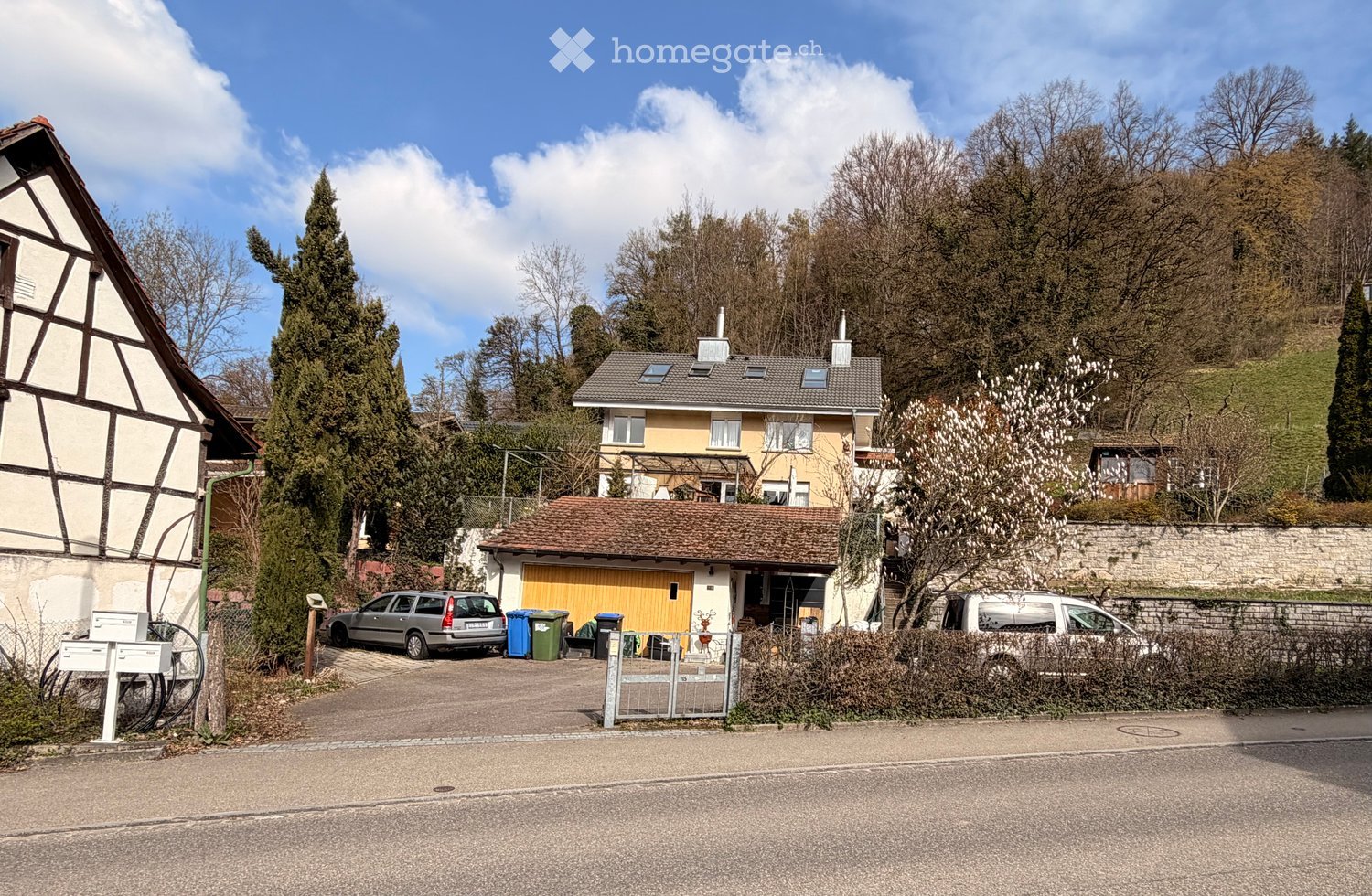 A two-story house with a yellow facade and a tiled roof, situated in a residential area with trees and a clear blue sky. The house features a garage with a wooden door and a driveway with parked cars.
