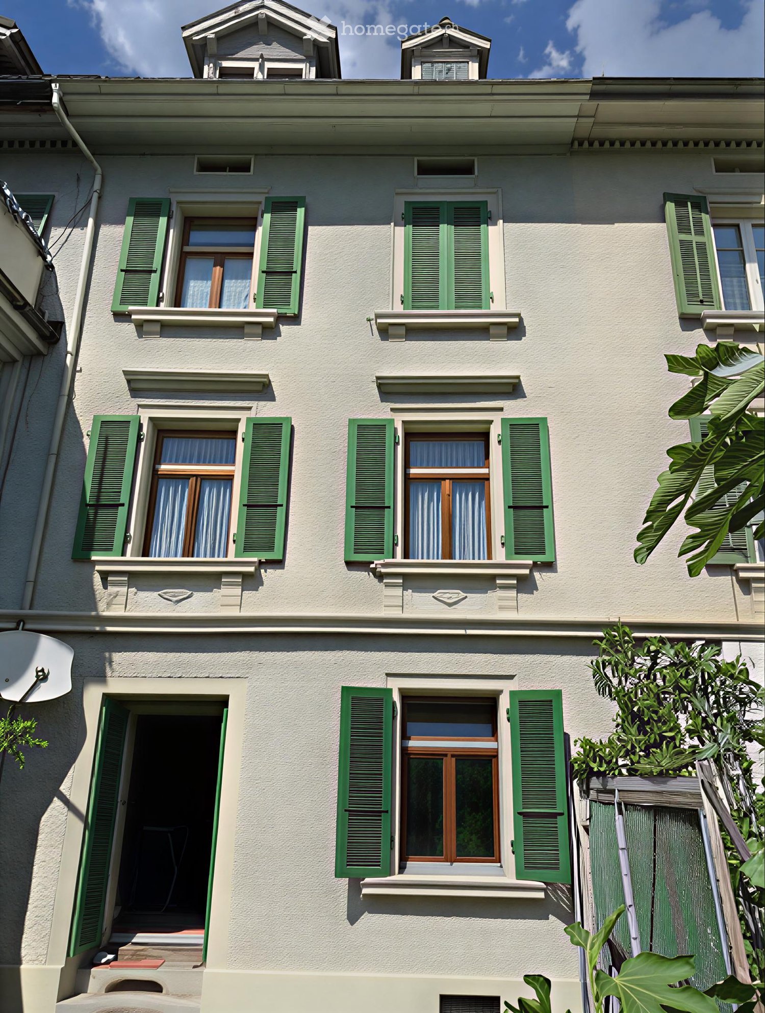 Three-story residential building with light-colored facade, green shutters, multiple windows, and a small balcony on the second floor.
