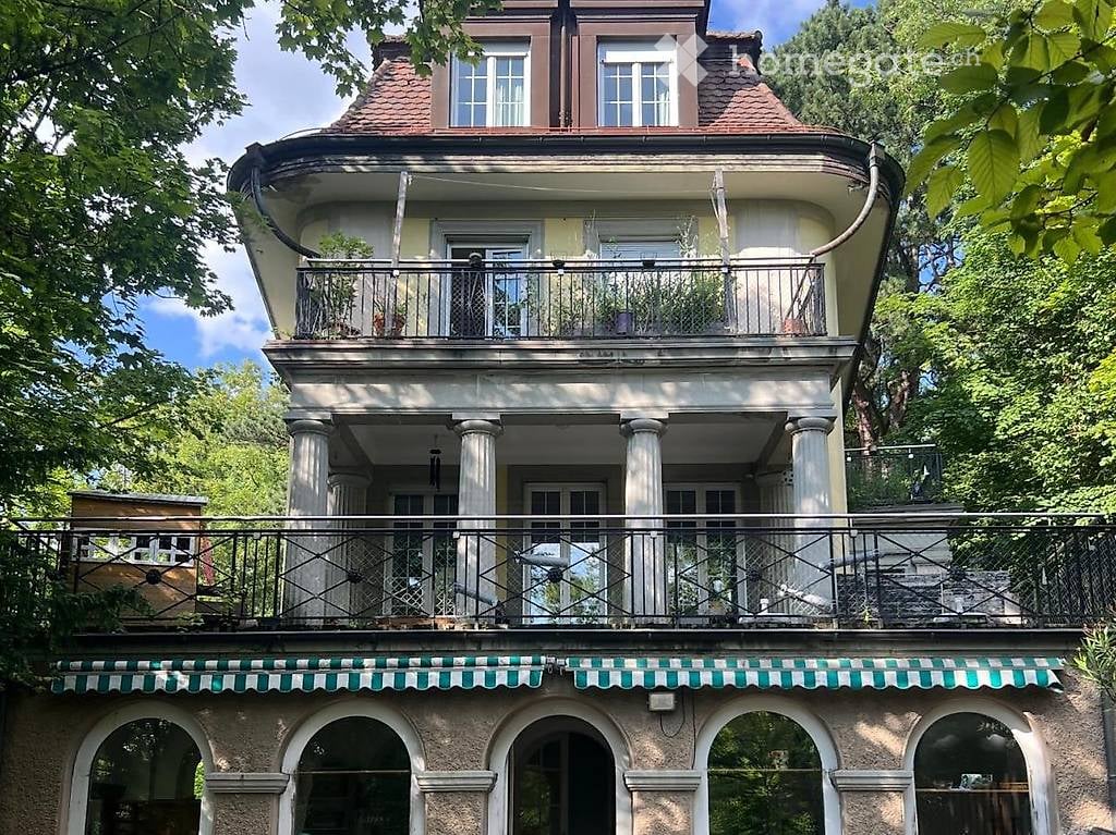 Building. A three-story building with a balcony and decorative columns, surrounded by greenery.