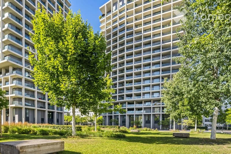 Modern high-rise residential building with multiple balconies and large windows, surrounded by green spaces and trees.