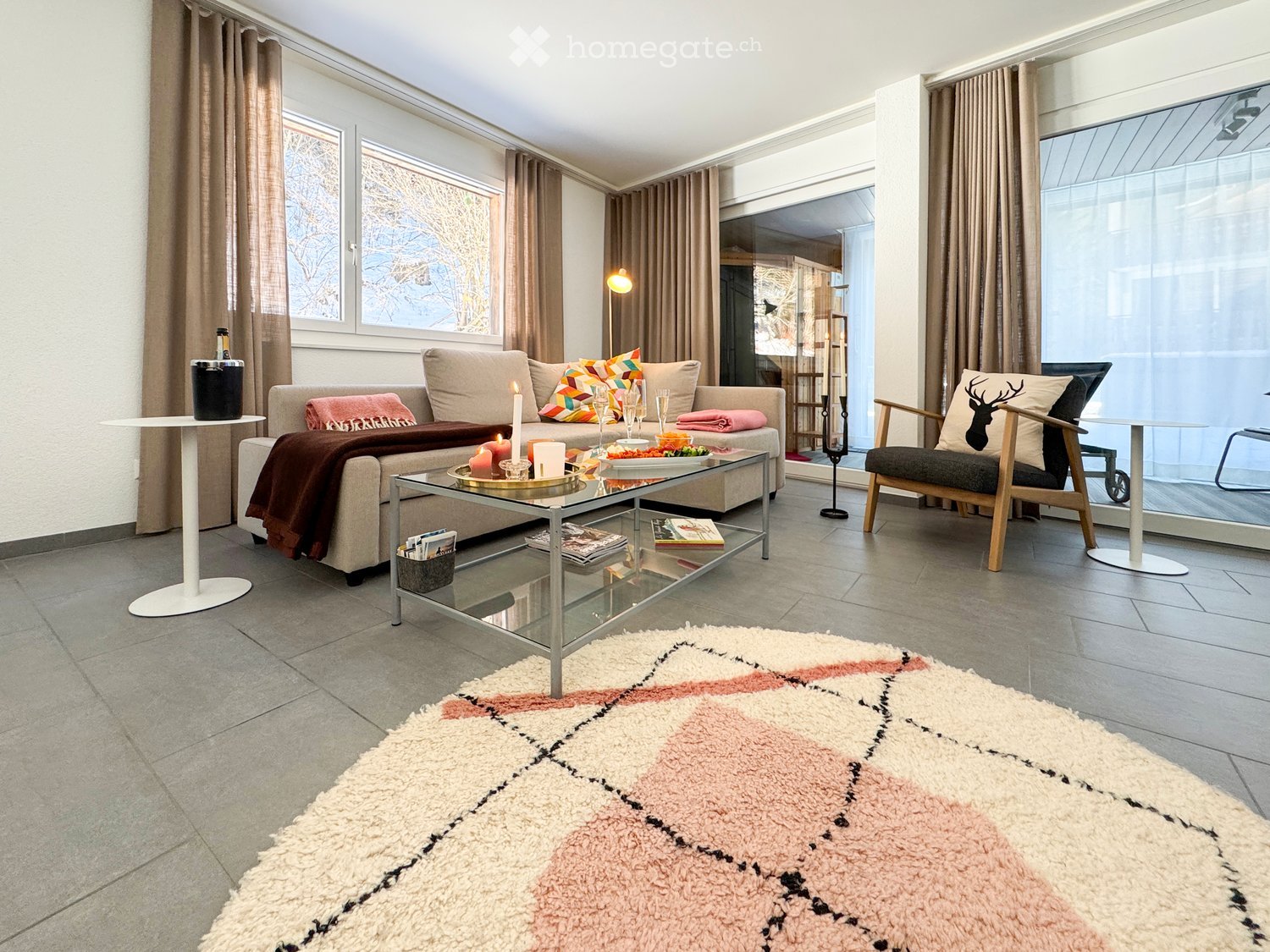 Modern living room with a beige sofa, glass coffee table, colorful cushions, a pink and white shag rug, and a wooden chair with a deer pillow.