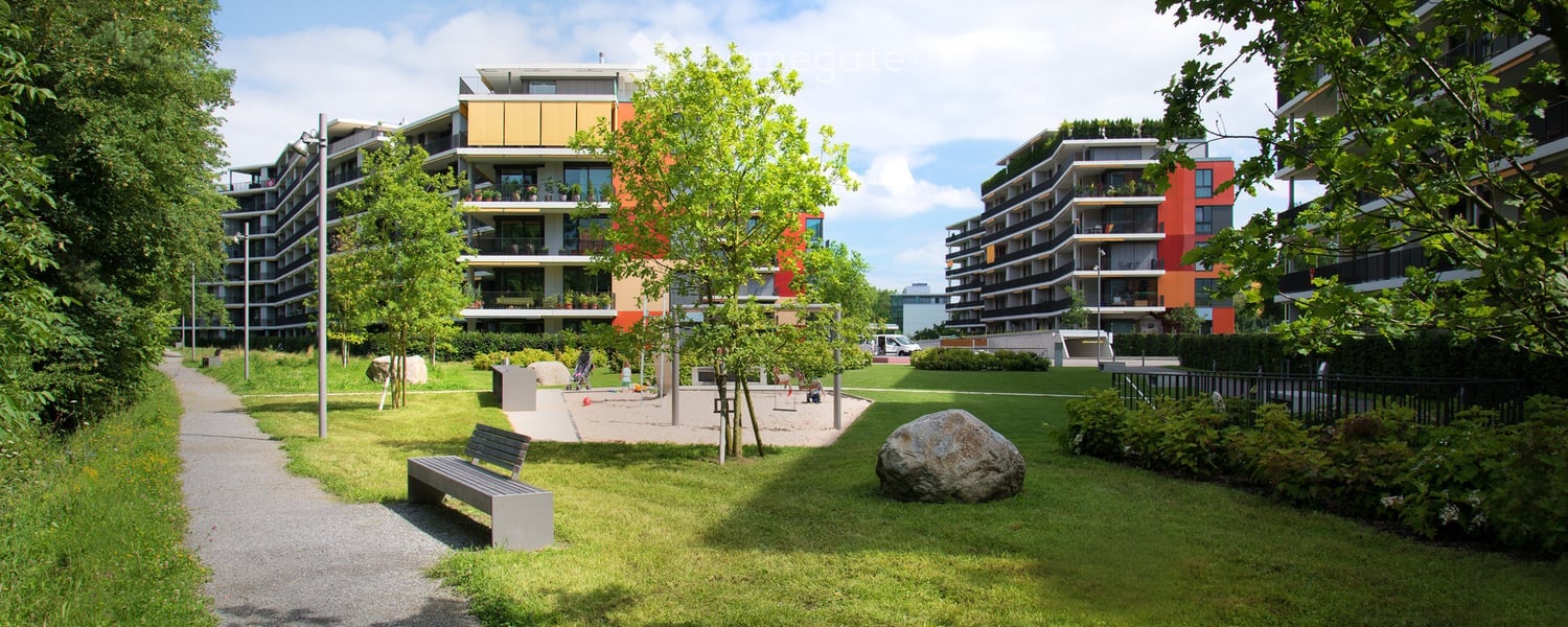 A well-maintained park area with a bench, trees, and a large rock, surrounded by modern apartment buildings.
